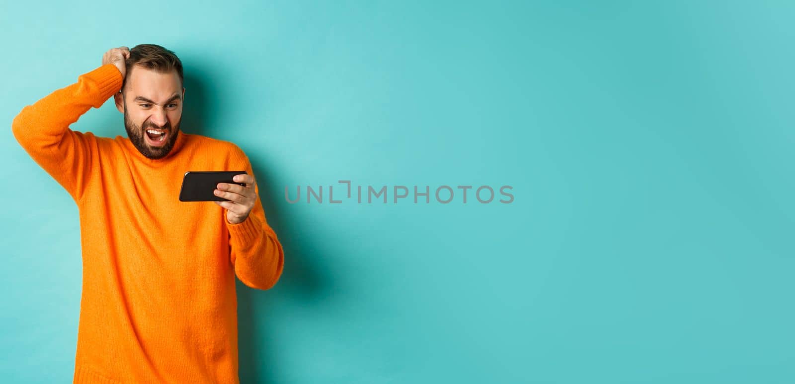 Image of man looking confused at mobile phone, watching something online, standing puzzled against light blue background. Copy space
