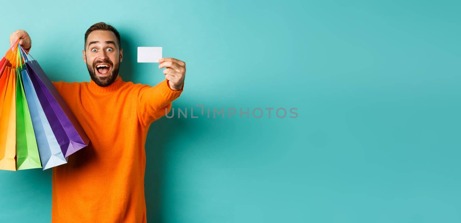 Happy aduly man showing credit card and shopping bags, standing against turquoise background.