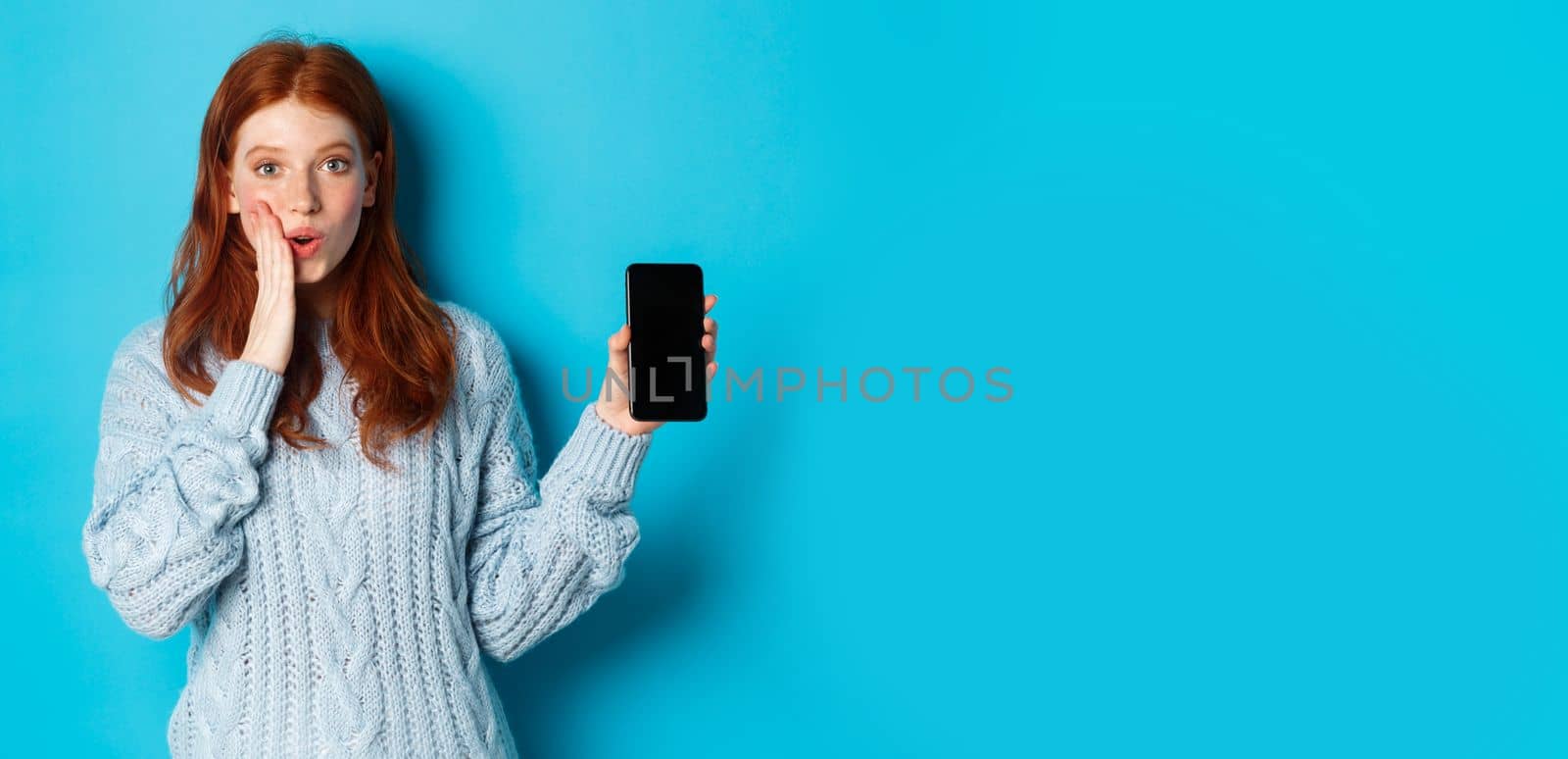 Amazed redhead girl looking at camera, showing smartphone screen, demonstrating online offer, standing over blue background.