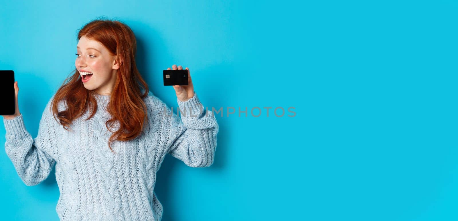 Excited redhead girl showing mobile phone screen and credit card, demonstrating online store or application, standing over blue background by Benzoix