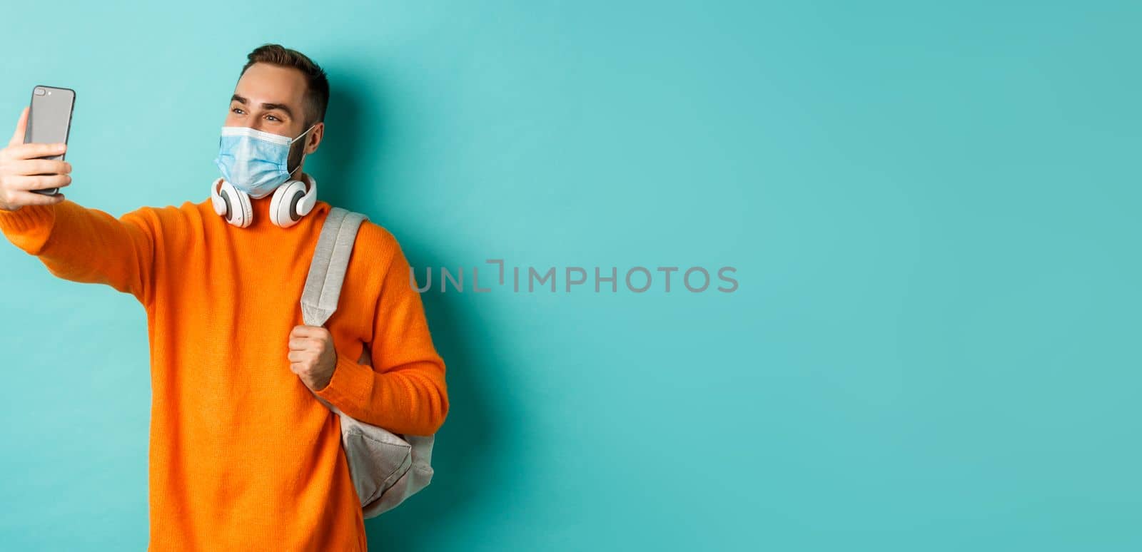 Young modern man with headphones and backpack, taking selfie on mobile phone in medical mask, standing over light blue background.