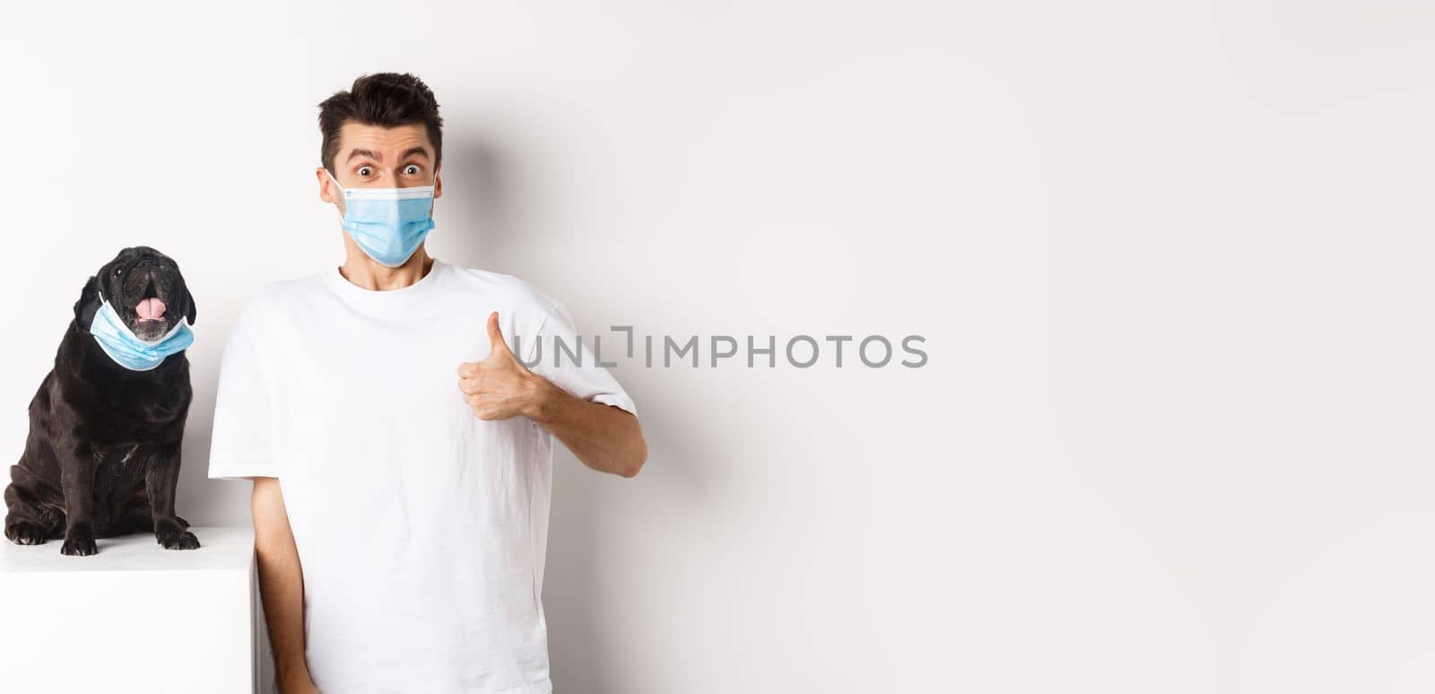 Covid-19, animals and quarantine concept. Image of funny young man and small dog in medical masks, owner showing thumb up in approval, praise something, white background by Benzoix