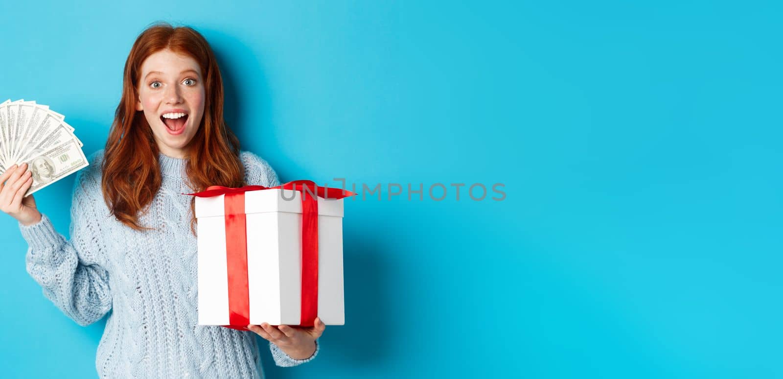 Christmas and shopping concept. Happy redhead woman holding money and big xmas present, showing dollars and gift, smiling pleased, standing over blue background.