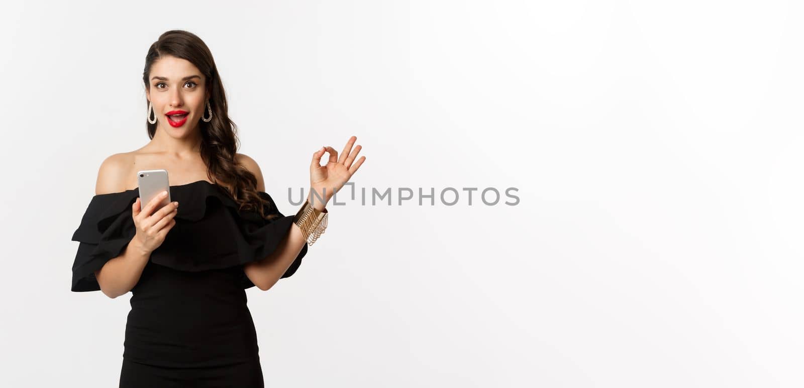 Online shopping concept. Attractive woman in trendy black dress, makeup, showing okay sign in approval and using mobile phone app, white background.