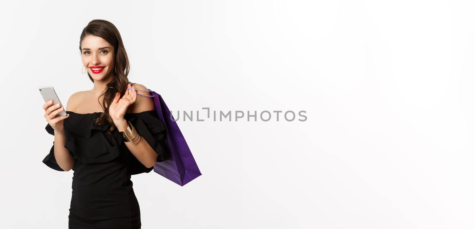 Beauty and shopping concept. Gorgeous woman in elegant black dress and makeup, order online on mobile phone, holding bag and smiling, standing over white background.