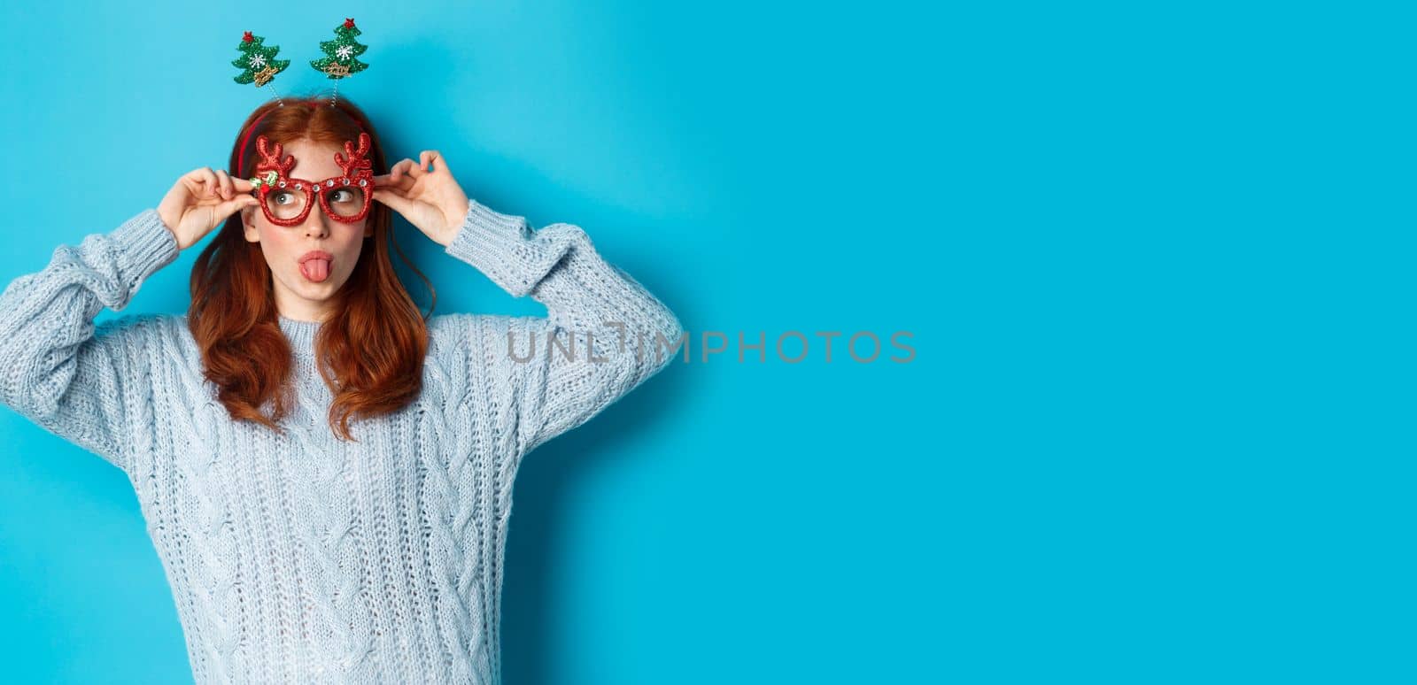 Christmas party and celebration concept. Silly redhead girl enjoying New Year, wearing funny glasses and headband, showing tongue and staring left at logo, blue background by Benzoix