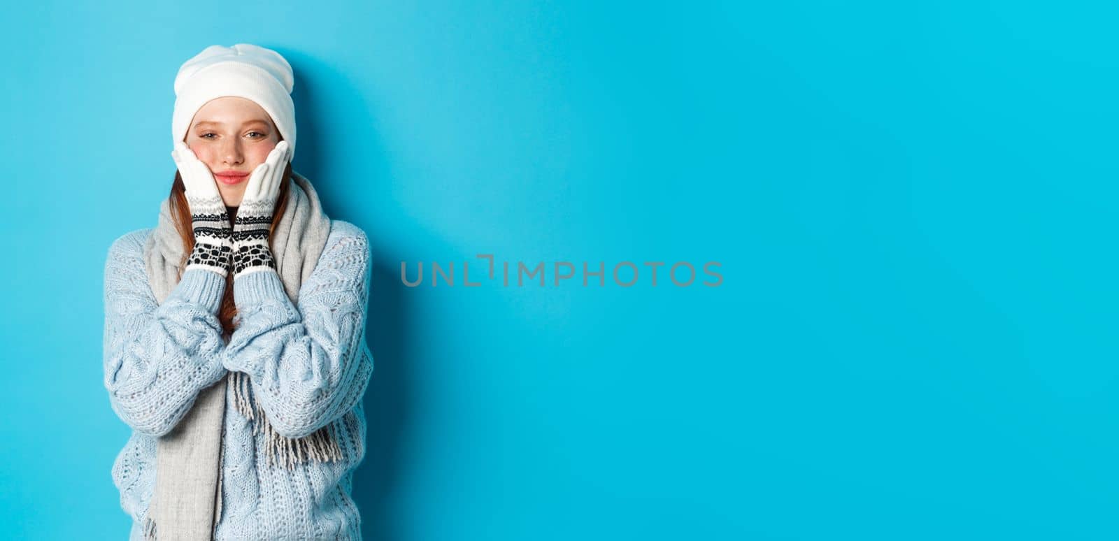 Winter and holidays concept. Cute girl in white beanie, sweater and gloves, squeezing cheeks and smiling pleased, warm-up after cold outdoors, standing over blue background by Benzoix