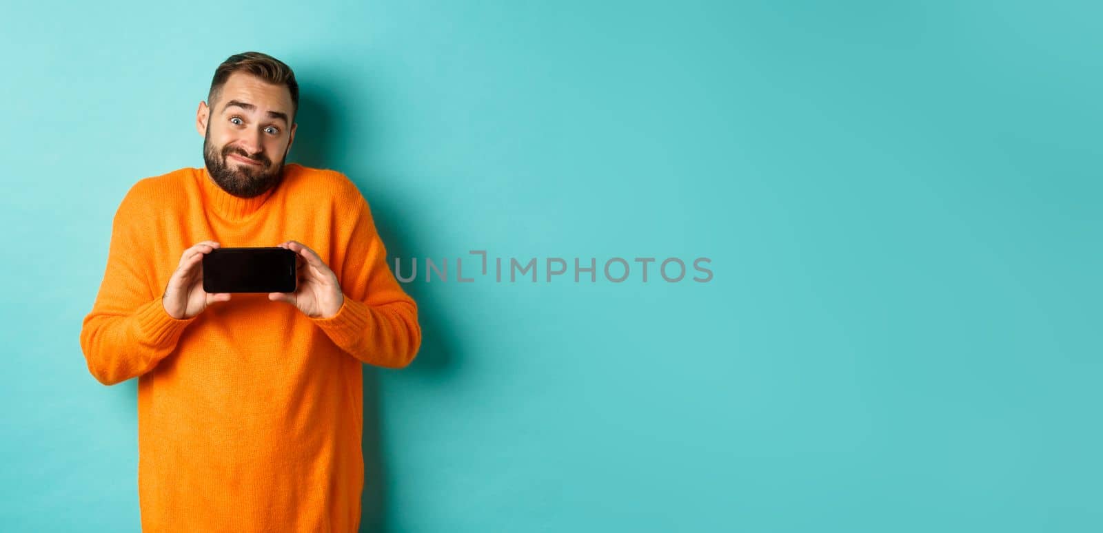 Clueless guy shrugging and showing mobile screen, indecisive emotion, standing in orange sweater over turquoise background.