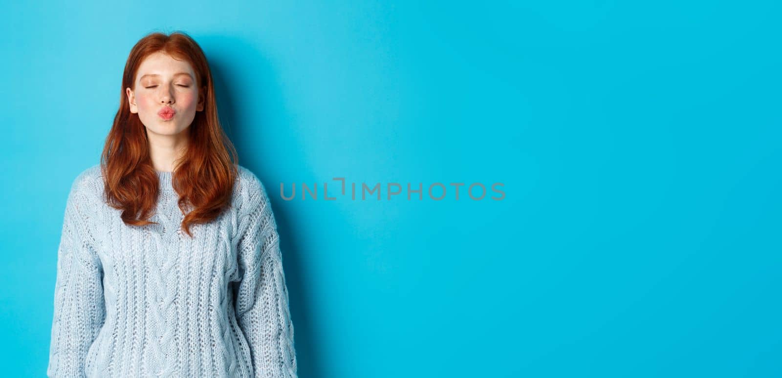 Cute redhead teen girl waiting for kiss, pucker lips and close eyes, standing in sweater against blue background.