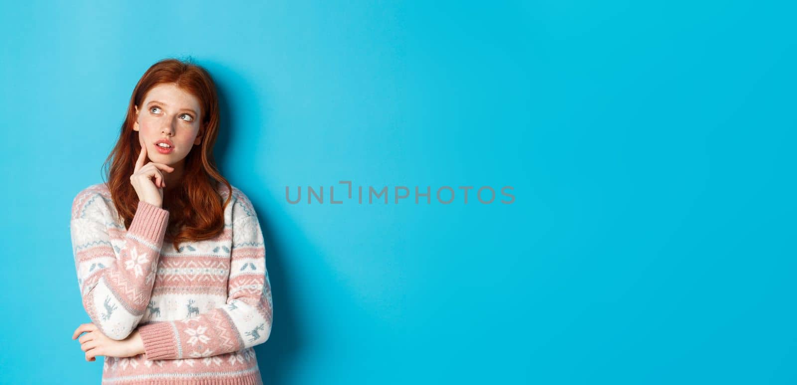 Image of thoughtful redhead girl looking at upper left corner, thinking or making choice, standing in winter sweater over blue background by Benzoix