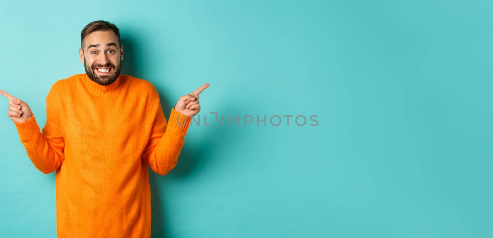 Confused handsome man shrugging indecisive, pointing fingers sideways, cant choose between two variants, standing against light blue background.