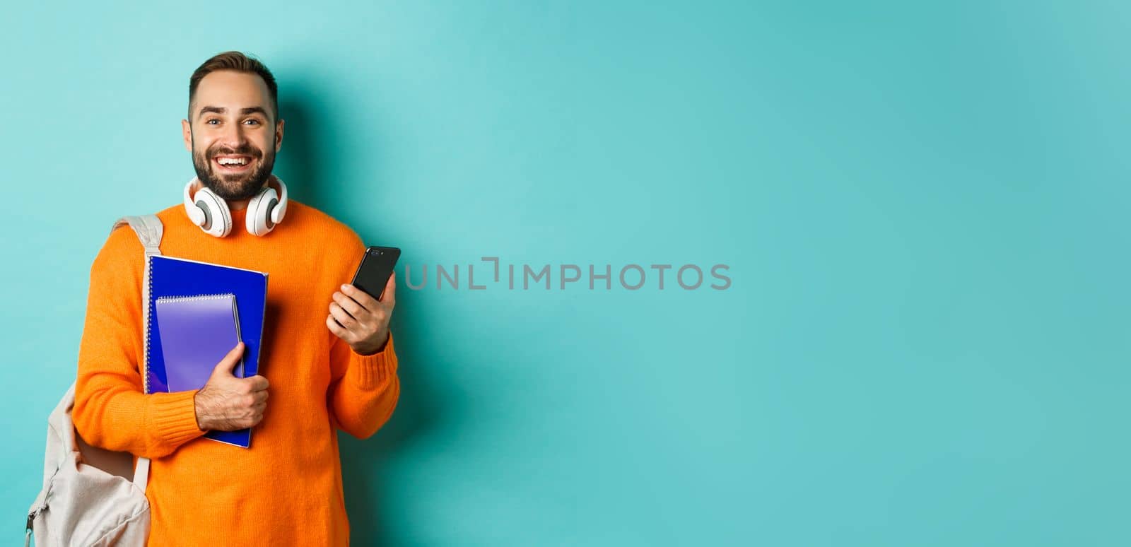 Education. Handsome male student with headphones and backpack, using smartphone and holding notebooks, smiling happy, standing over turquoise background.