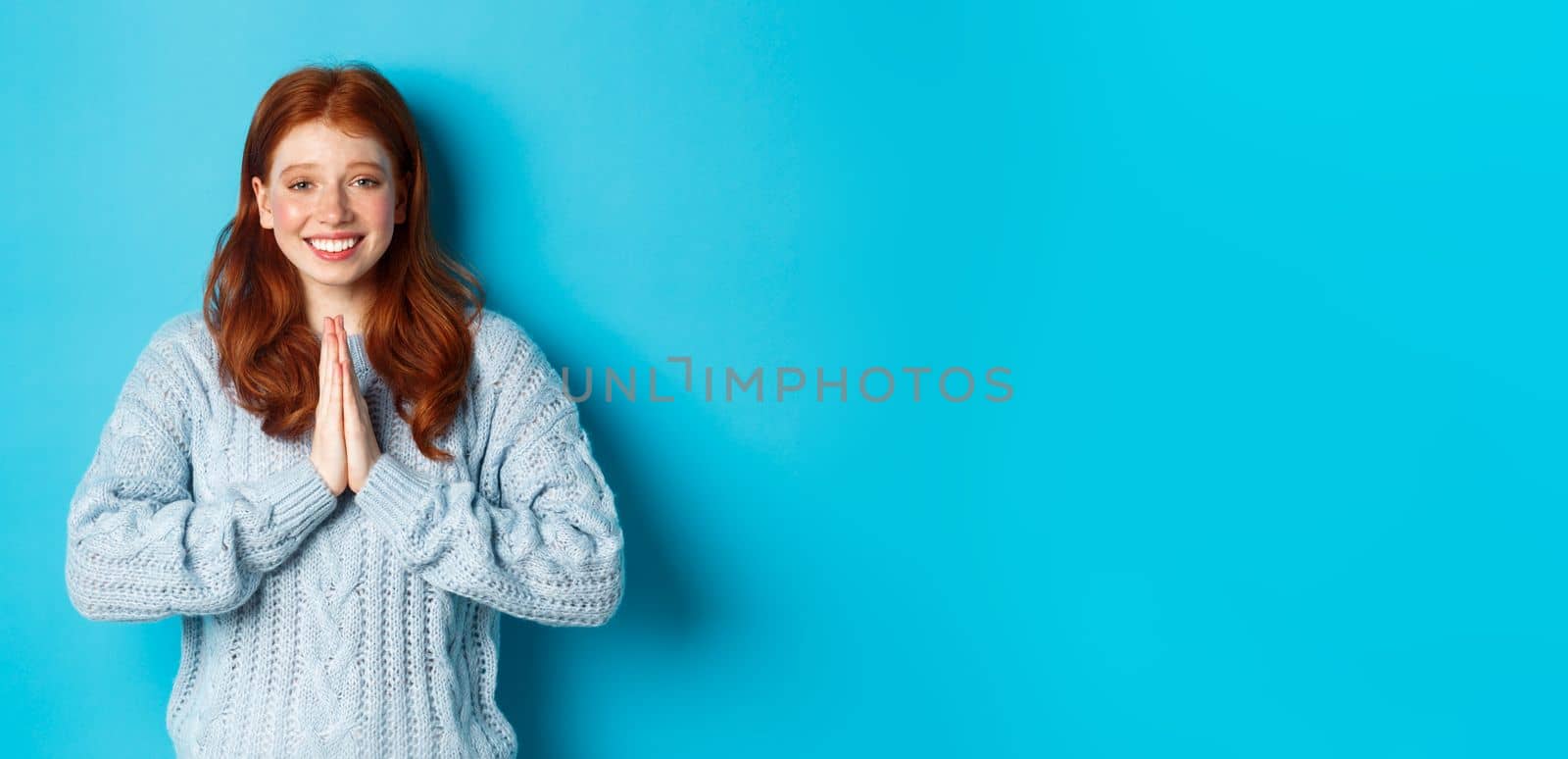 Cute redhead girl saying thank you, smiling and looking at camera, expressing gratitude, standing against blue background.