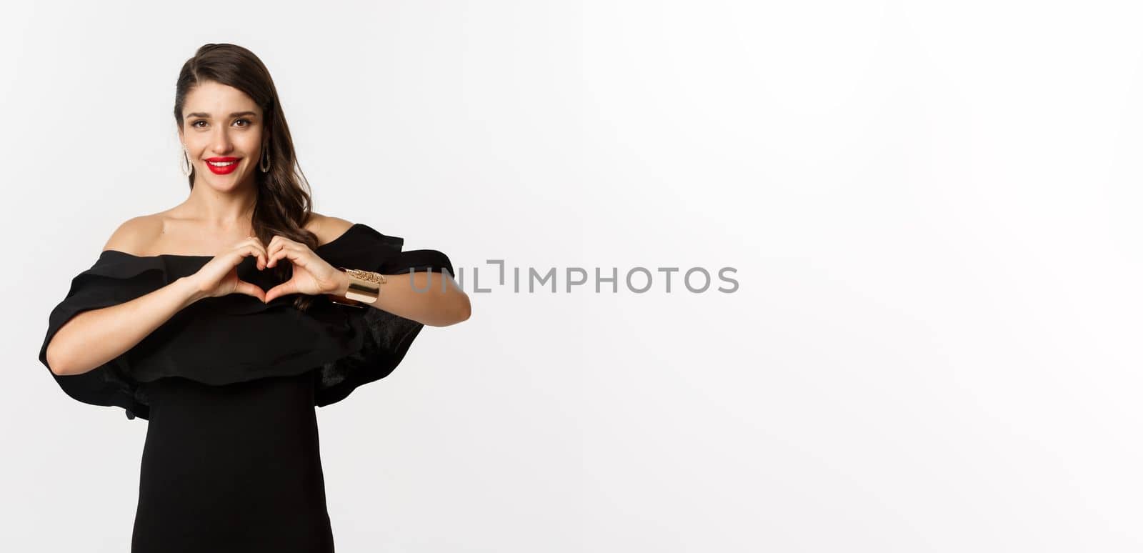 Fashion and beauty. Beautiful young bruentte female showing heart sign and smiling, express love and sympathy, wearing black dress, white background.