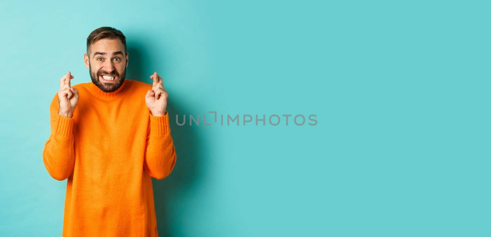 Worried man making a wish, hoping for something, cross fingers good luck, standing nervous over turquoise background.