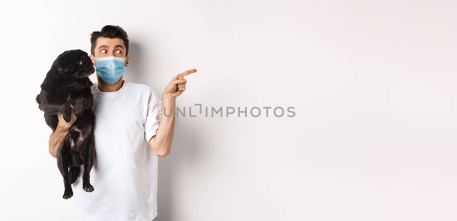 Covid-19, animals and quarantine concept. Young man in face mask holding cute black pug, dog and pet owner looking right at copy space, standing over white background.