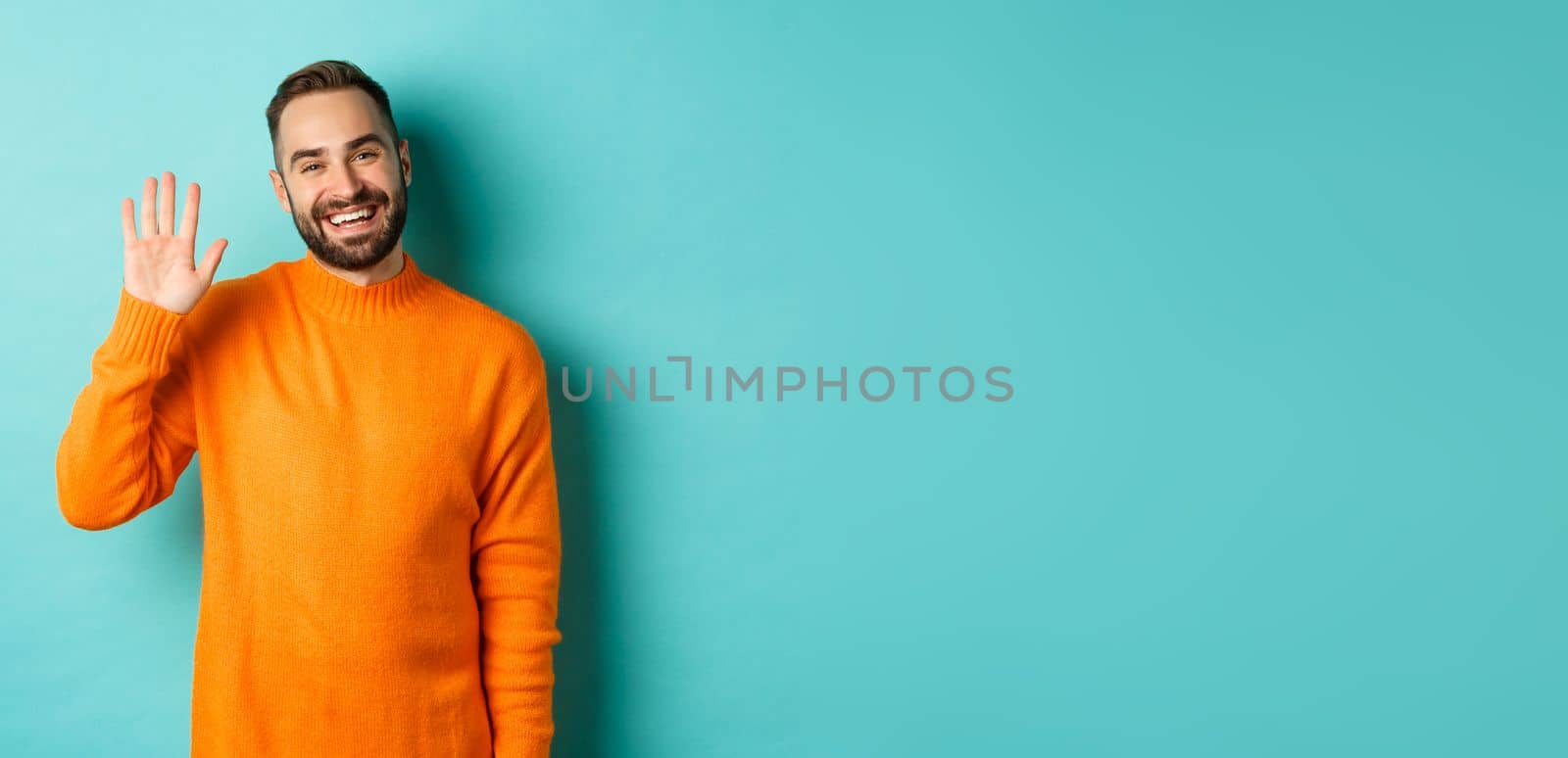 Photo of friendly young man saying hello, smiling and waiving hand, greeting you, standing in orange sweater over light blue background by Benzoix