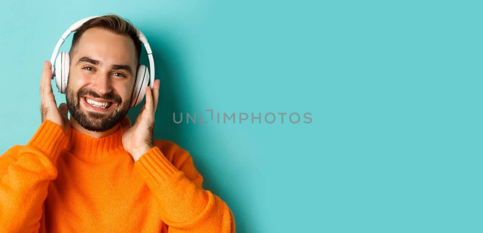 Close-up of handsome modern man listening music in headphones, standing in orange sweater over turquoise background by Benzoix