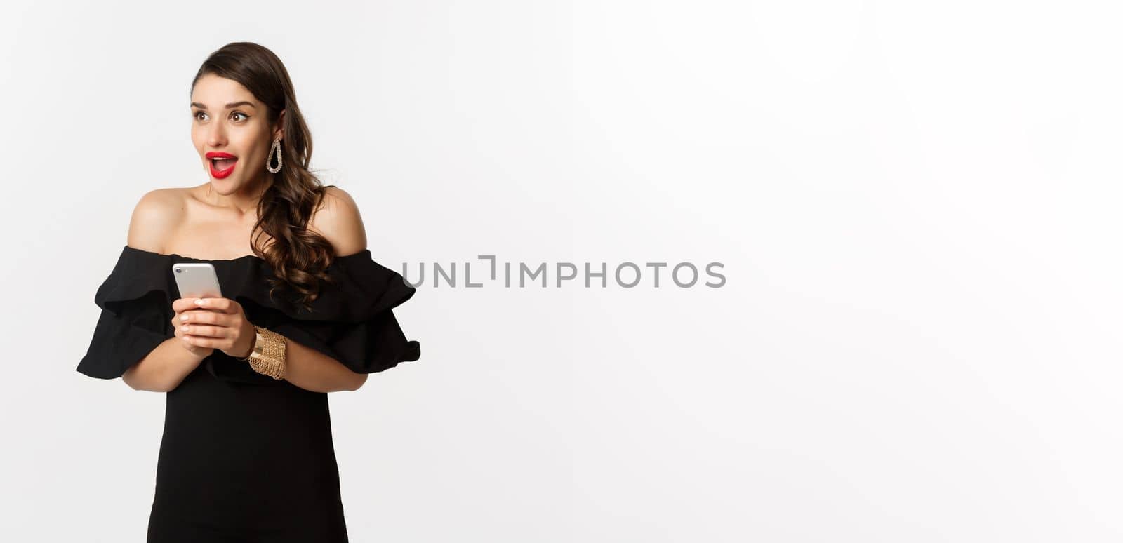Image of excited woman in black glamour dress, red lipstick, using mobile phone and looking left amazed, standing over white background by Benzoix