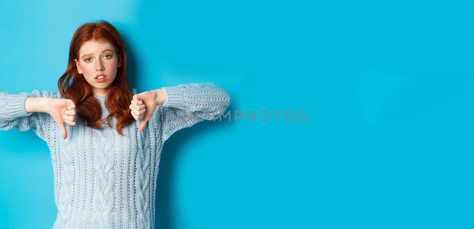 Bored and skeptical redhead girl showing thumbs down, looking unamused and uninterested, standing over blue background by Benzoix