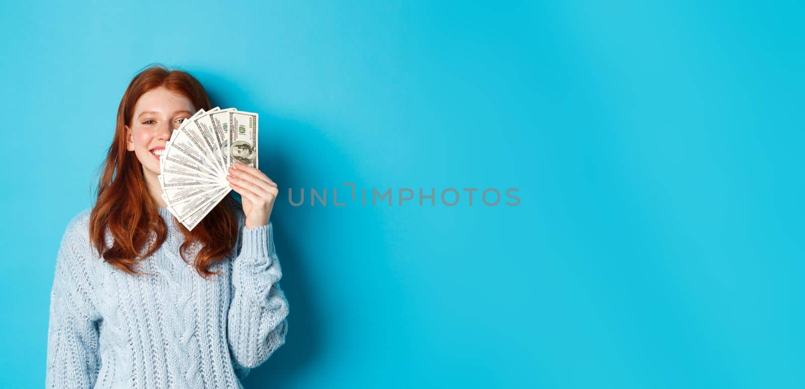 Delighted redhead woman showing money near face, smiling satisfied, standing ready for shopping over blue background by Benzoix