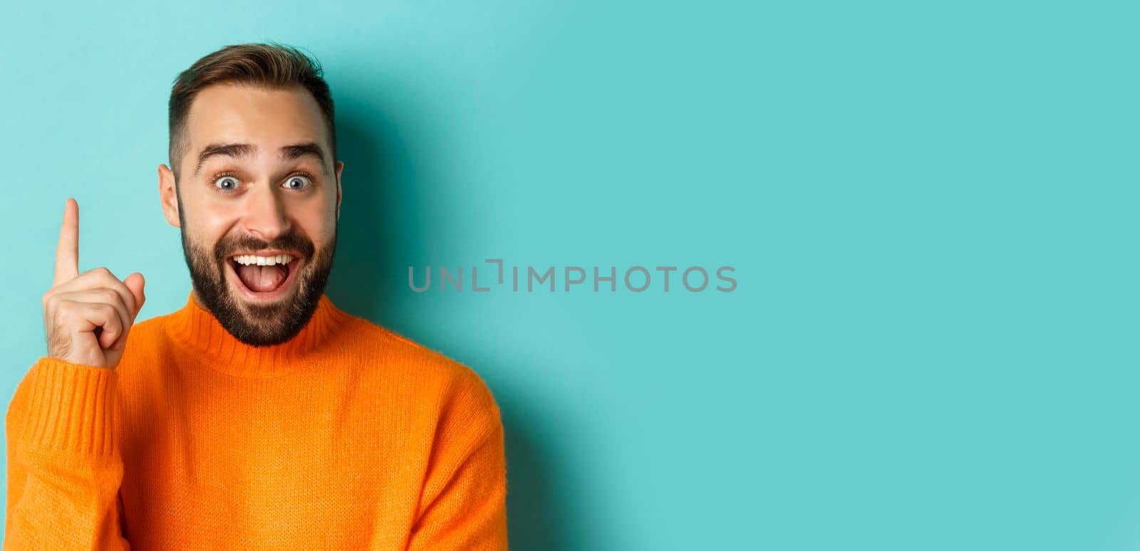 Close-up of handsome man having an idea, raising finger up and smiling excited, standing in orange sweater.