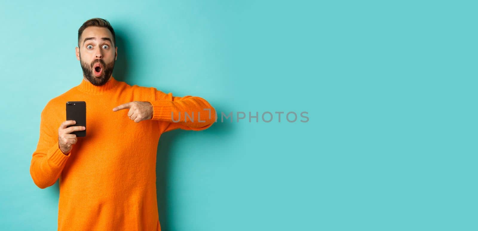 Image of impressed man showing something on mobile phone, pointing at smartphone and gasping amazed, standing in orange sweater over light blue background by Benzoix