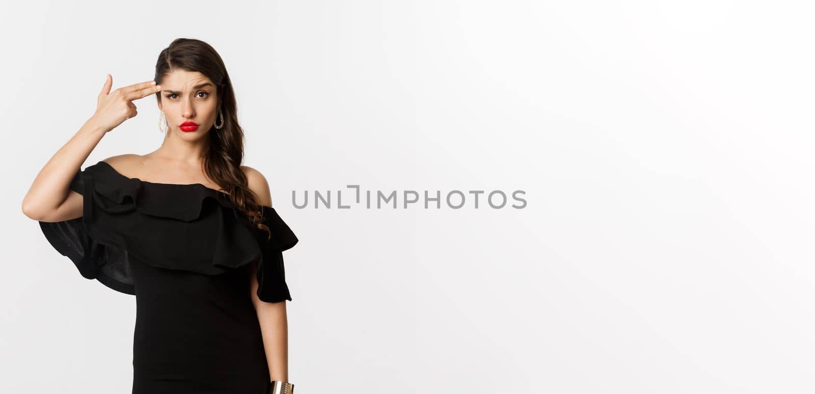 Fashion and beauty. Annoyed young woman in black dress making finger gun gesture near head, shooting herself from irritation, standing over white background.