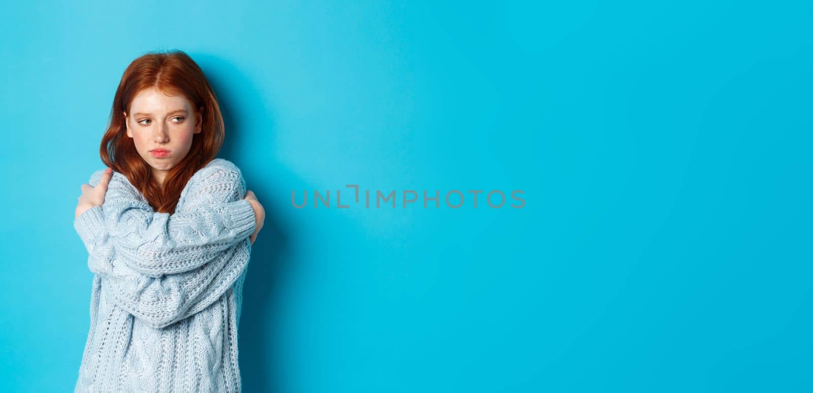 Silly and cute redhead girl pucker lips and looking offended, hugging herself and looking away offended, sulking and being upset, standing over blue background by Benzoix