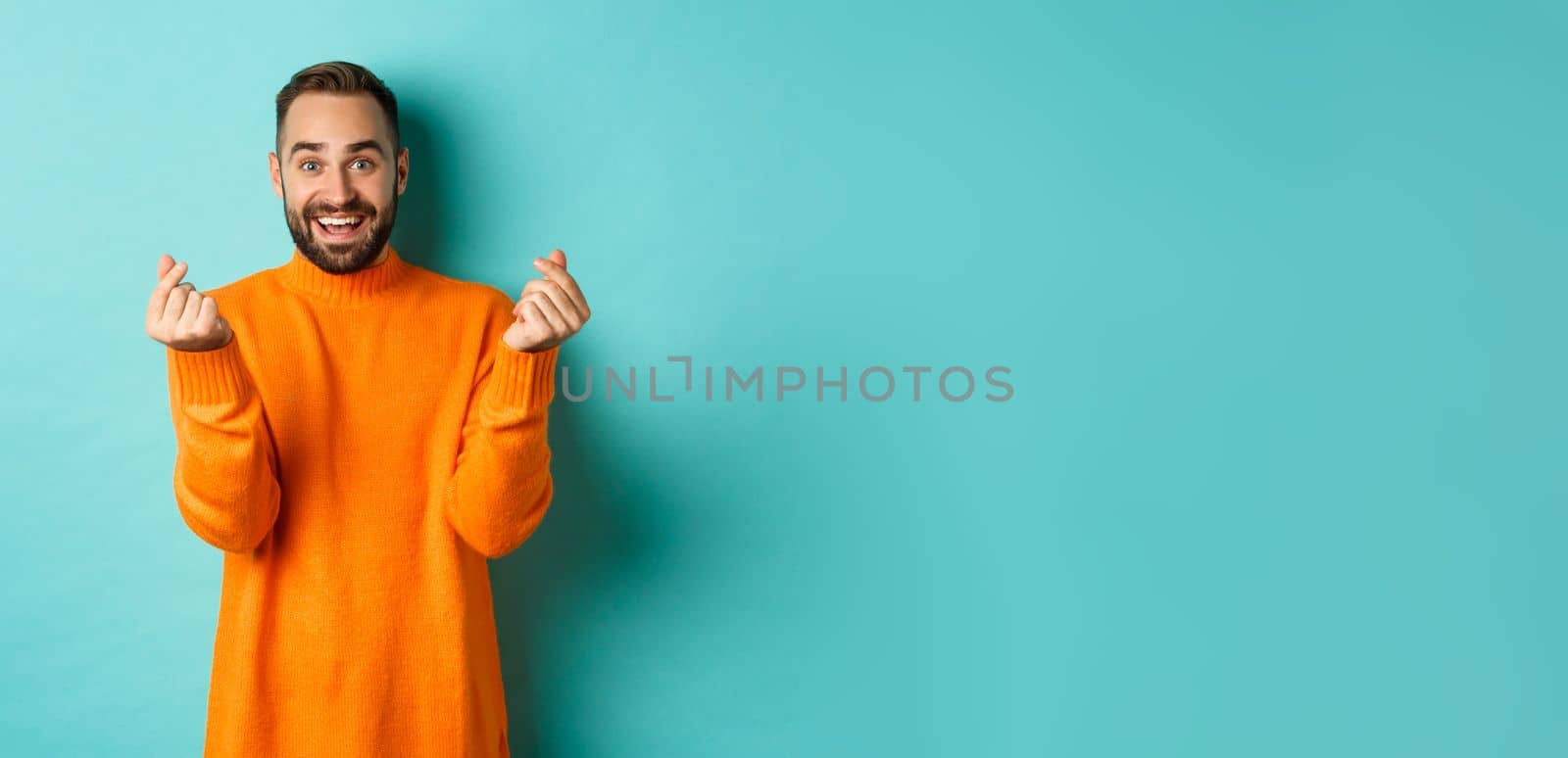 Smiling handsome man showing hearts and looking happy, lucky gesture, standing in orange sweater over light blue background.