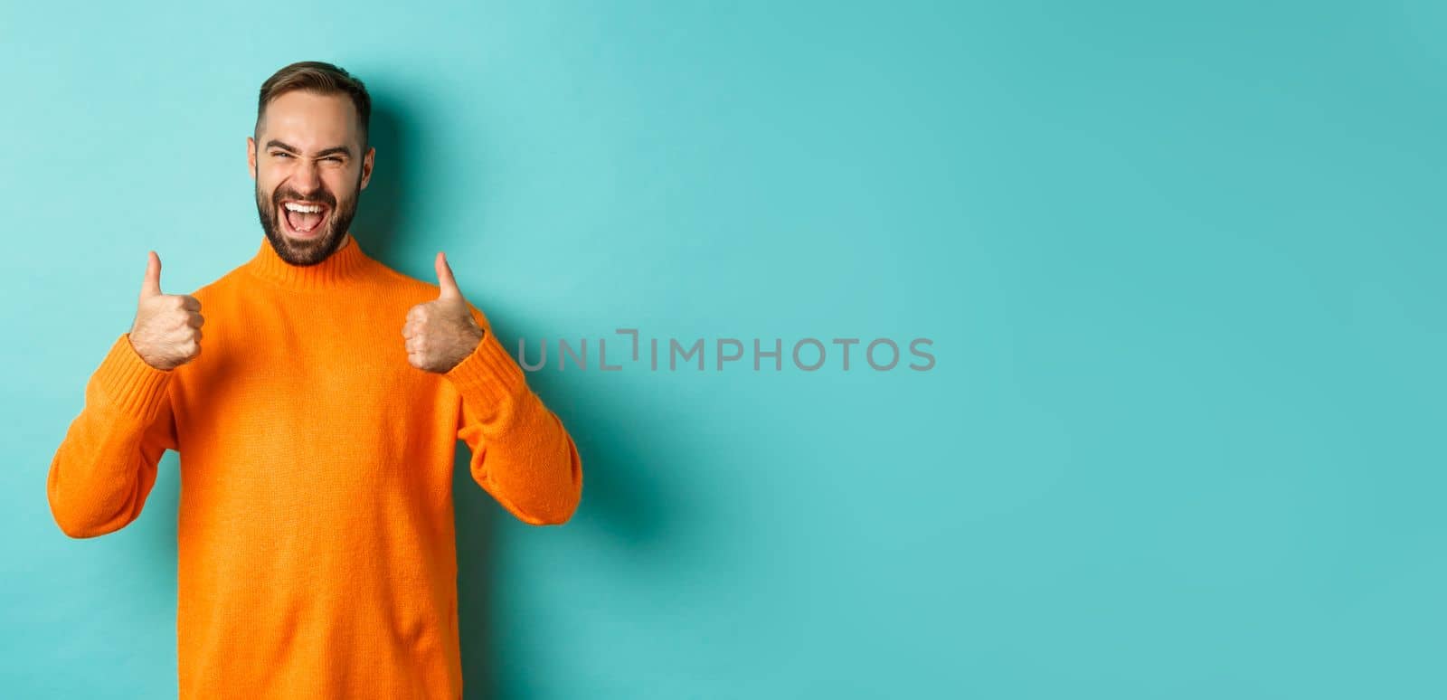 Super good. Satisfied happy man showing thumbs up, agree with you, praise excellent work, looking pleased, standing in orange sweater against light blue background by Benzoix