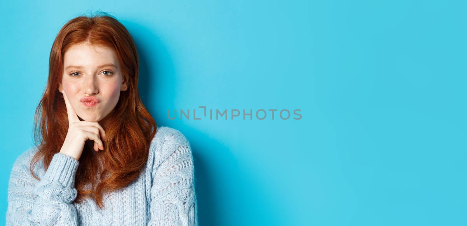 Close-up of beautiful redhead girl thinking, pucker lips and staring thoughtfula t camera, making choice, standing over blue background by Benzoix