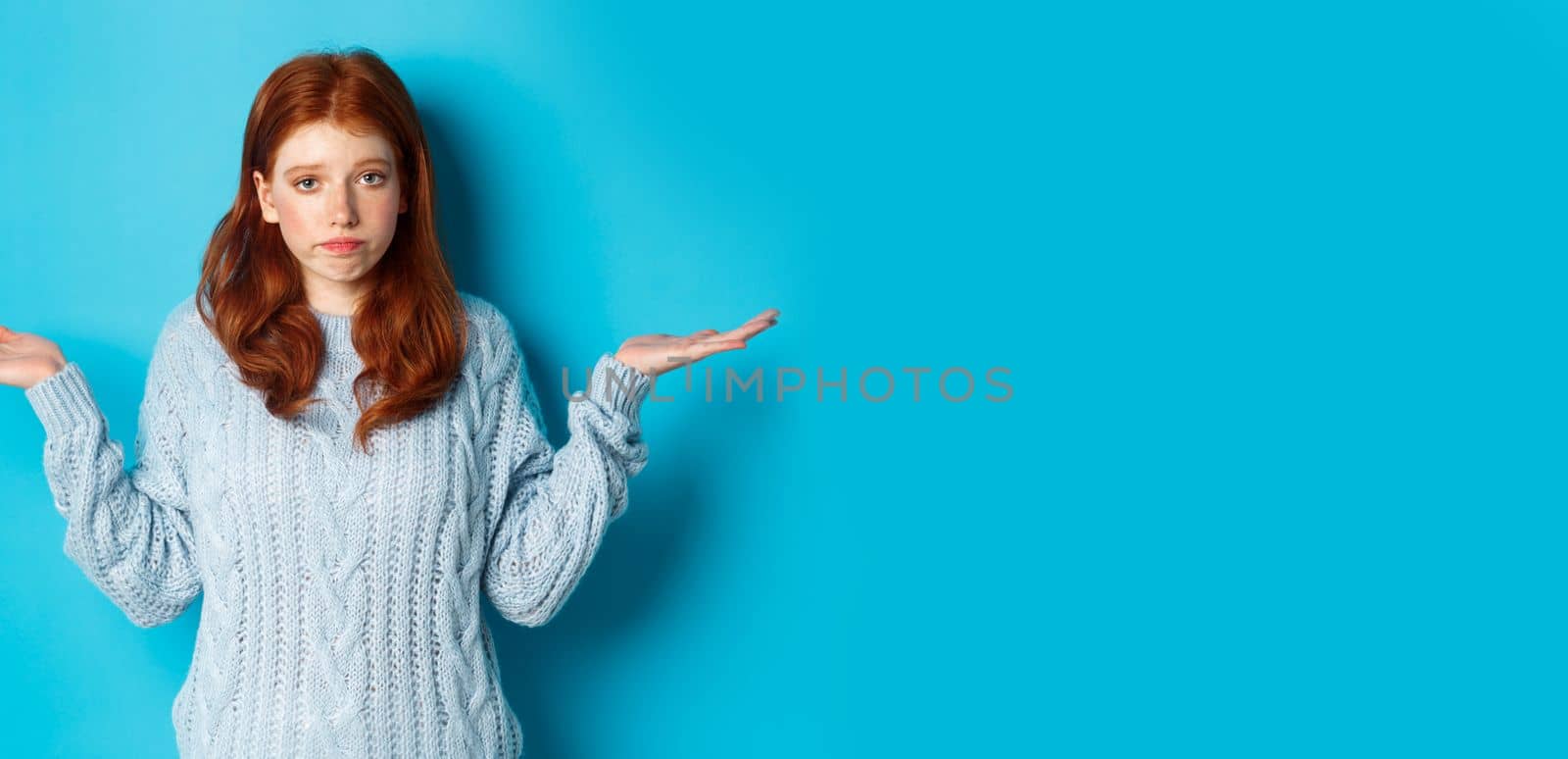 Clueless redhead girl shrugging and saying sorry, standing puzzled against blue background, have no idea.
