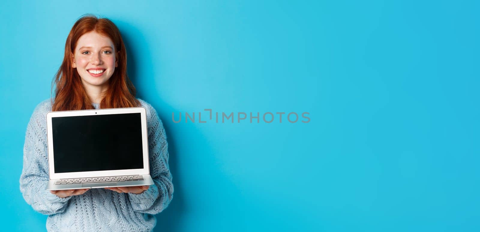 Young smiling woman with red hair and freckles showing computer screen, holding laptop and demonstrate online promo, standing over blue background by Benzoix