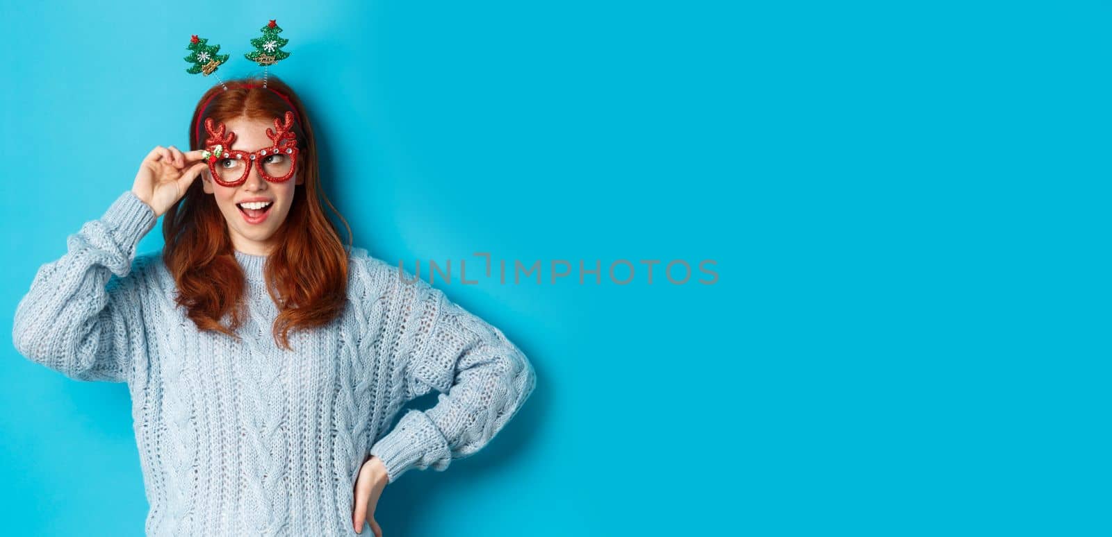 Christmas party and celebration concept. Cute redhead teen girl celebrating New Year, wearing xmas tree headband and funny glasses, looking left amused, blue background.