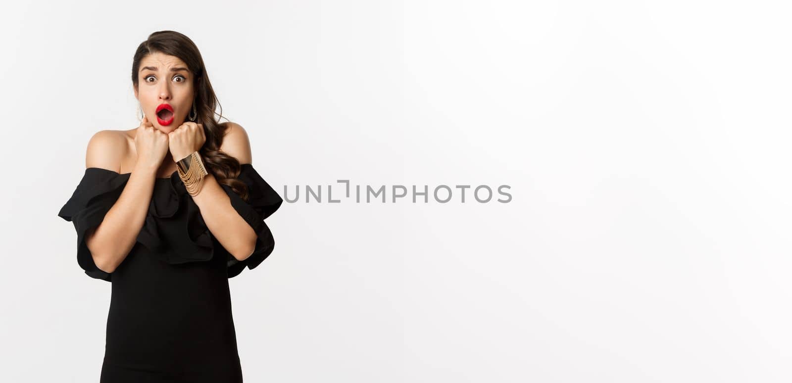 Portrait of glamour woman looking scared and shocked at camera, staring at something with fear, standing in black dress against white background.