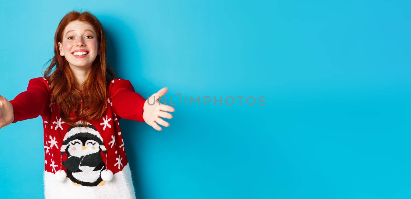 Winter holidays and Christmas Eve concept. Beautiful redhead girl in xmas sweater reaching for hug, extend hands for cuddles and smiling, standing in sweater against blue background by Benzoix