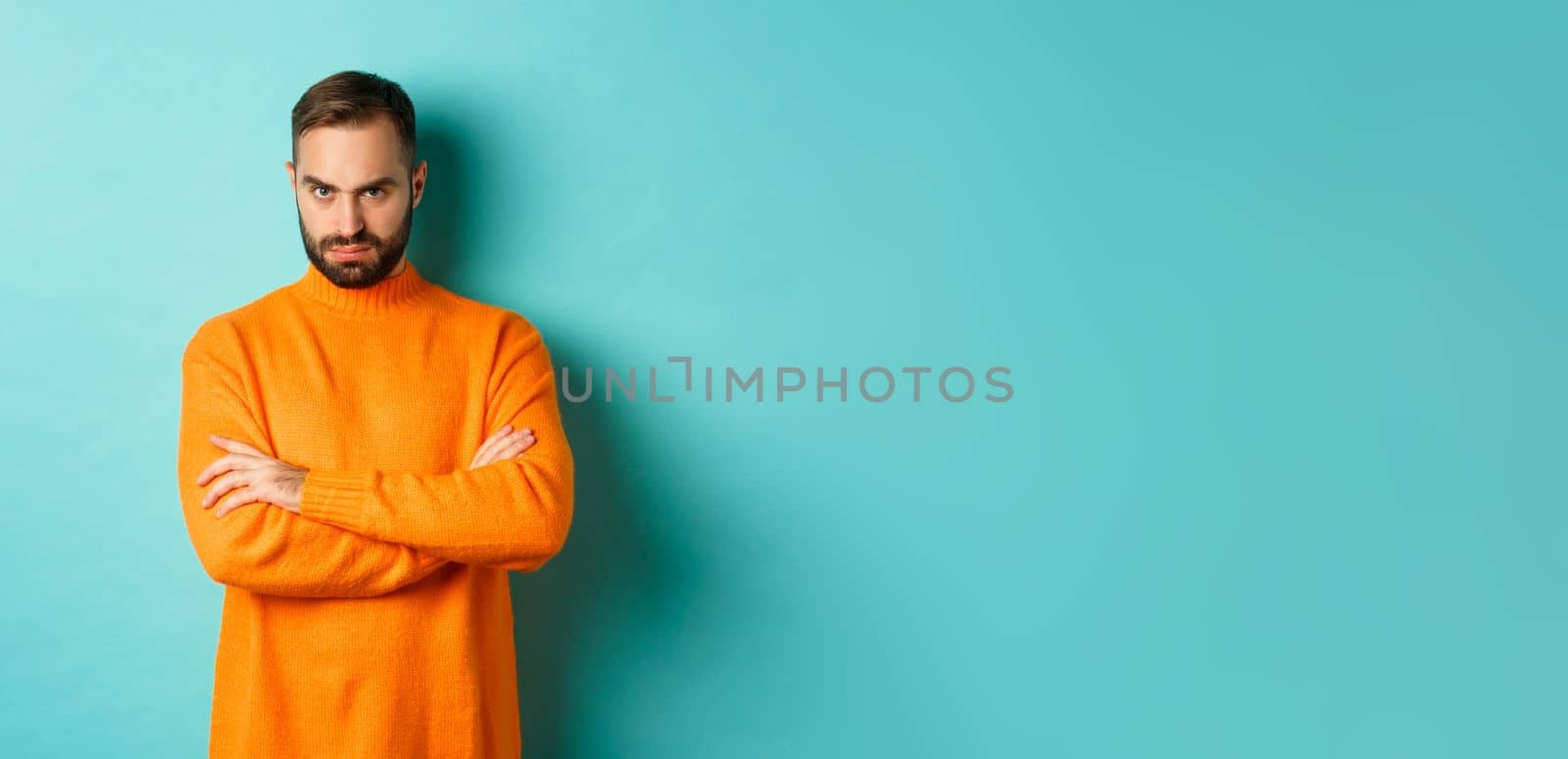 Offended man looking angry at you, cross arms on chest and stare mad, standing in orange sweater against turquoise background.