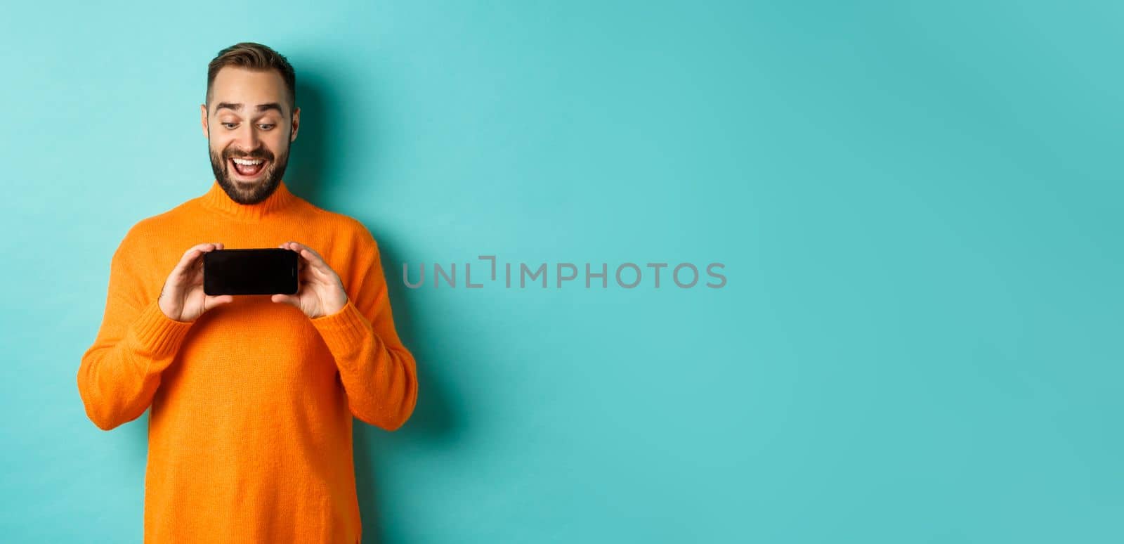 Excited guy demonstrating online store, showing mobile screen and looking amazed, standing over light blue background.