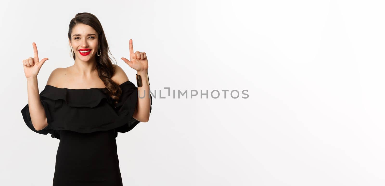 Fashion and beauty. Charming woman with red lips, black dress, smiling happy and pointing fingers up, showing logo, white background by Benzoix