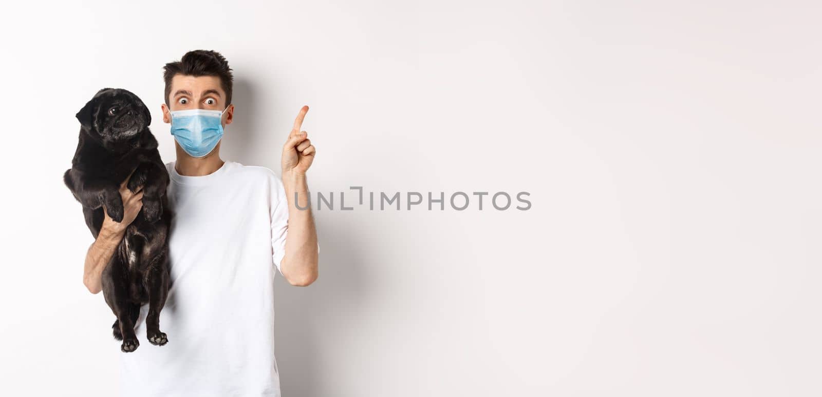 Covid-19, animals and quarantine concept. Young man in medical mask holding cute black pug, dog looking right and owner pointing at logo, white background.