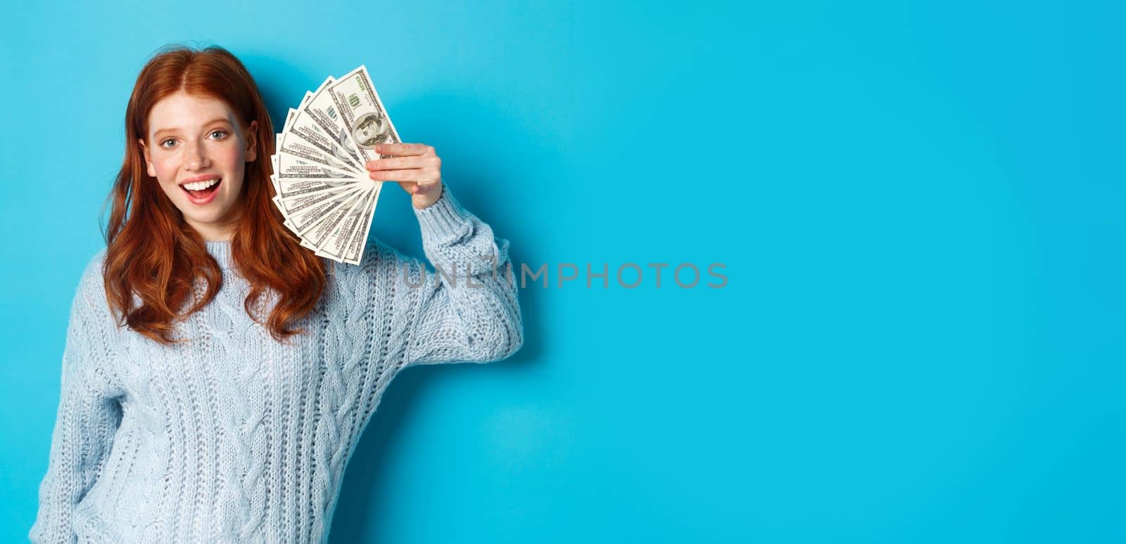 Cheerful redhead woman in sweater showing dollars, smiling pleased and holding money, standing over blue background.