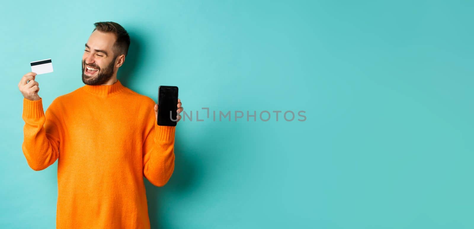 Online shopping. Satisfied man using credit card and showing mobile screen, looking pleased, standing over light blue background.