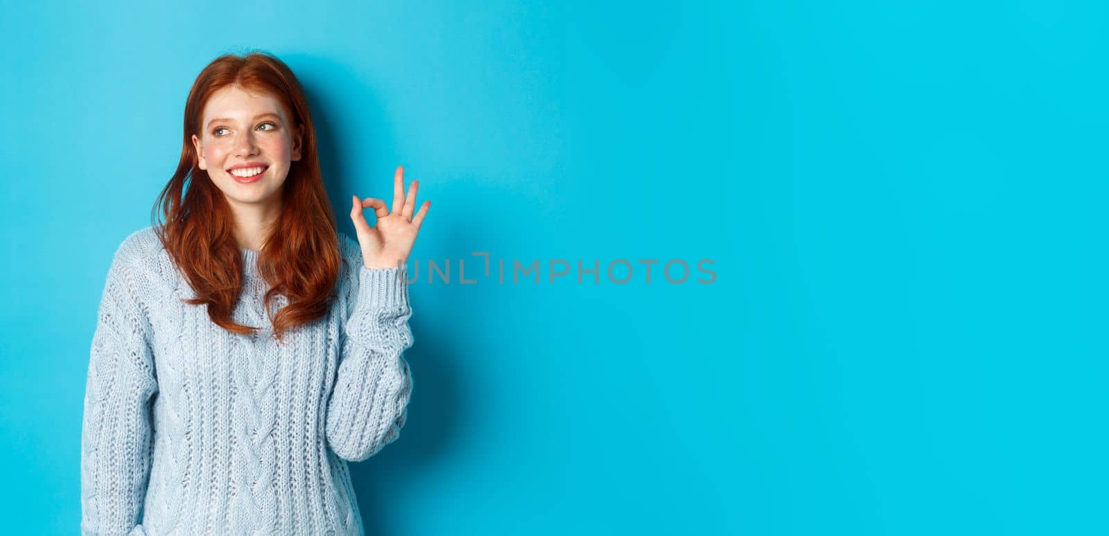 Image of beautiful redhead woman showing okay sign and smiling satisfied, like and agree, standing against blue background.