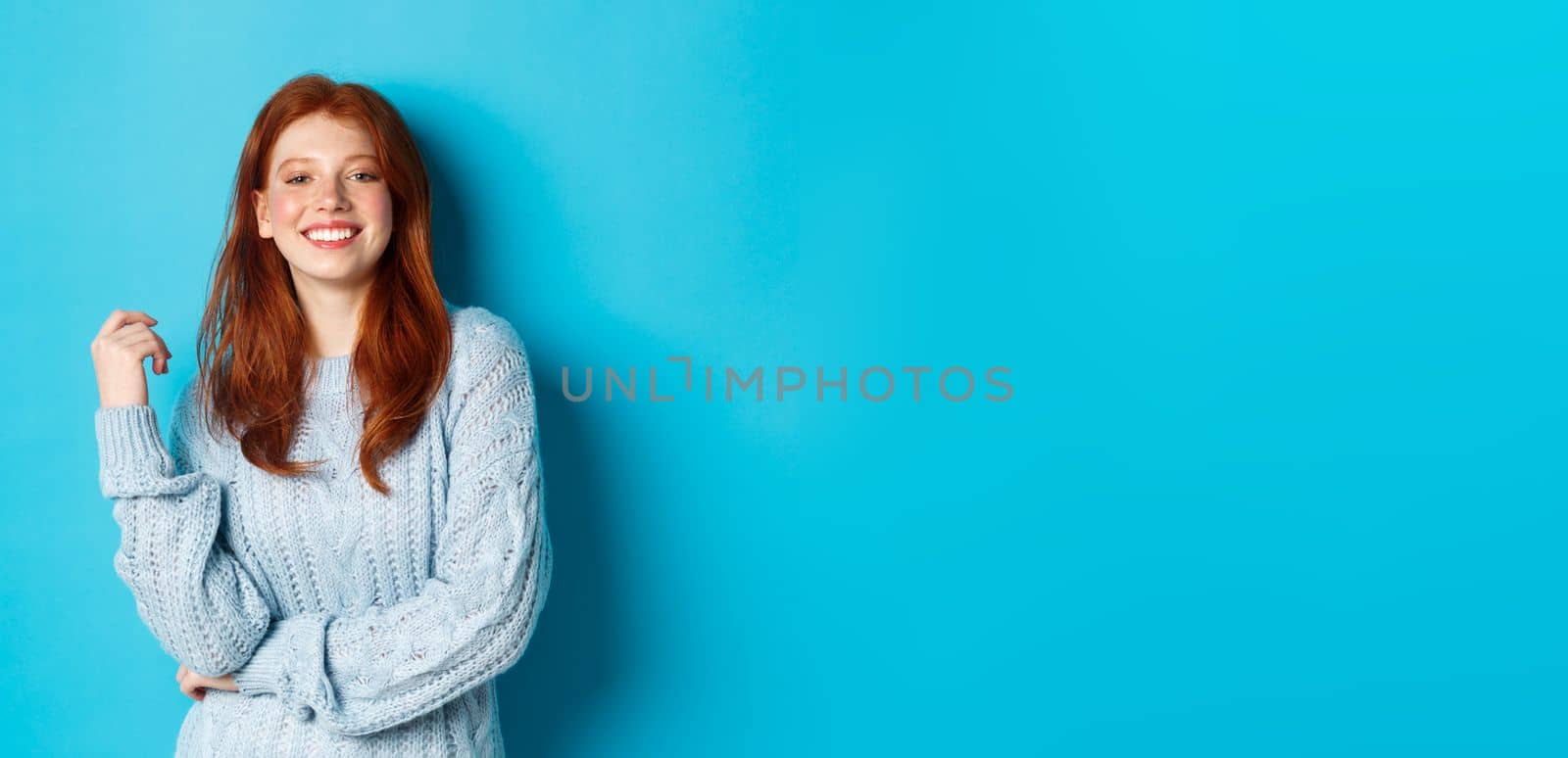 Happy redhead female in sweater, looking pleased at camera and smiling, standing against blue background.