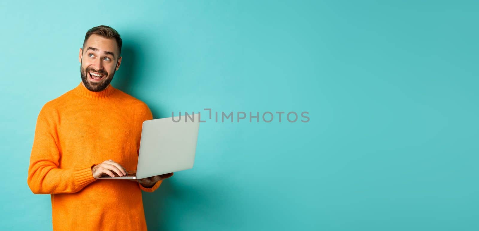 Handsome man doing online shopping, looking up thoughtful while using laptop, standing over light blue background. Copy space