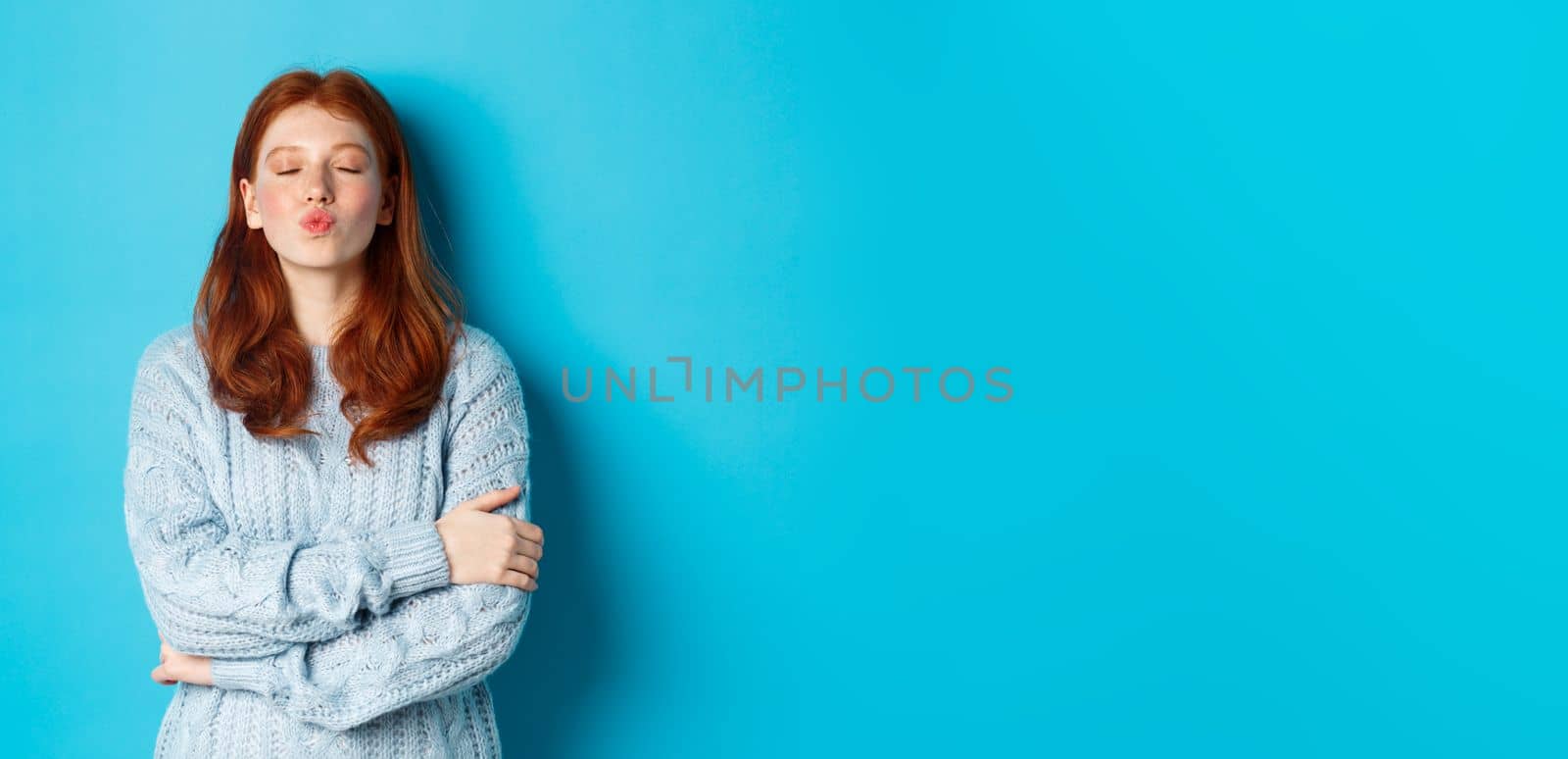 Cute redhead teen girl waiting for kiss, pucker lips and close eyes, standing in sweater against blue background.