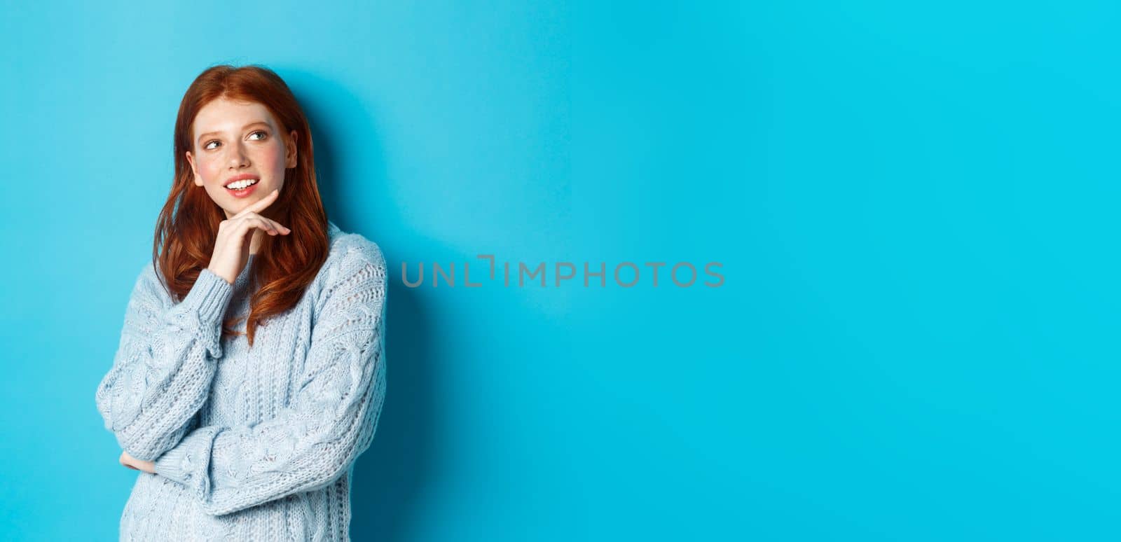 Thoughtful teenage girl with red hair, looking upper right corner logo and thinking, imaging something, standing over blue background.