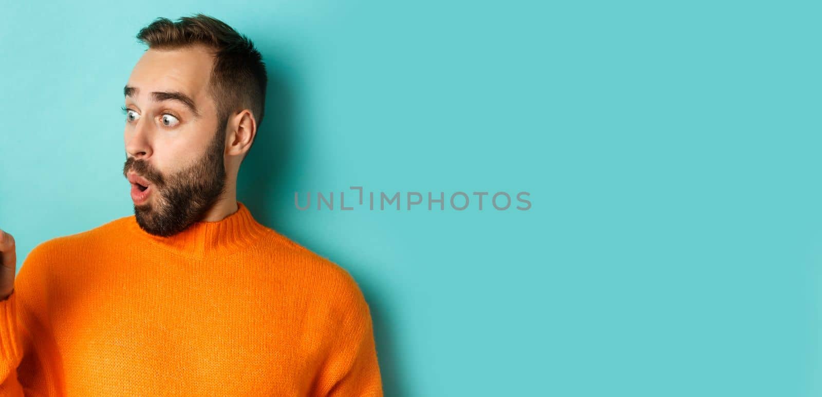 Close-up of young bearded man showing phone screen and looking amazed, wearing orange sweater, standing against studio background by Benzoix