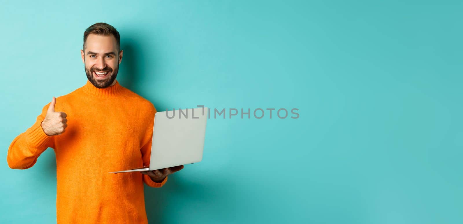Freelance and technology concept. Lucky man in orange sweater, showing thumb up while working with laptop, standing satisfied over light blue background.
